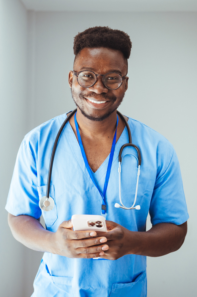 male nurse holding phone in blue scrubs with stethescope around neck smiles at camera